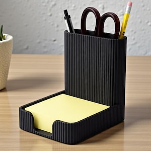 Pu&ograve; includere: Un organizer da scrivania nero con un design a coste, contenente una penna, forbici e una matita. Un blocco di post-it gialli si trova nella parte anteriore. L'organizer &egrave; su una superficie marrone chiaro.
