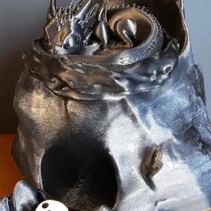 K&ouml;nnte beinhalten: Eine metallisch silberne Drachenfigur, die sich in einer h&ouml;hlenartigen Struktur zusammenrollt. Der Drache hat detaillierte Schuppen und H&ouml;rner. Ein wei&szlig;er W&uuml;rfel mit schwarzen Punkten liegt vor der H&ouml;hle. Fantasy-Dekorationsartikel.