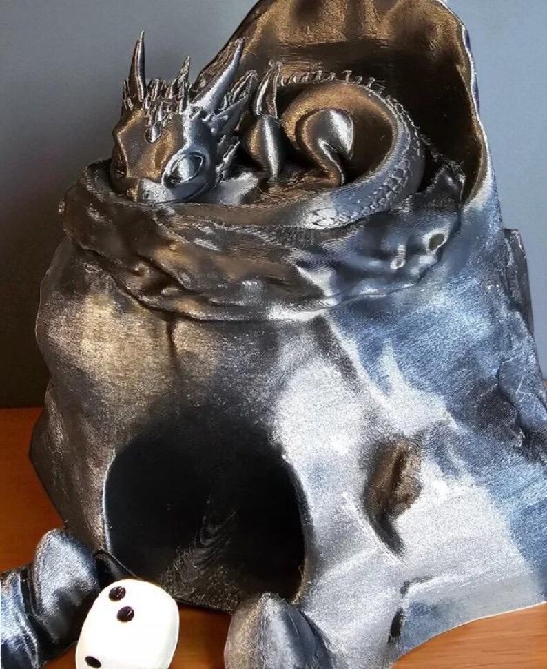 K&ouml;nnte beinhalten: Eine metallisch silberne Drachenfigur, die sich in einer h&ouml;hlenartigen Struktur zusammenrollt. Der Drache hat detaillierte Schuppen und H&ouml;rner. Ein wei&szlig;er W&uuml;rfel mit schwarzen Punkten liegt vor der H&ouml;hle. Fantasy-Dekorationsartikel.