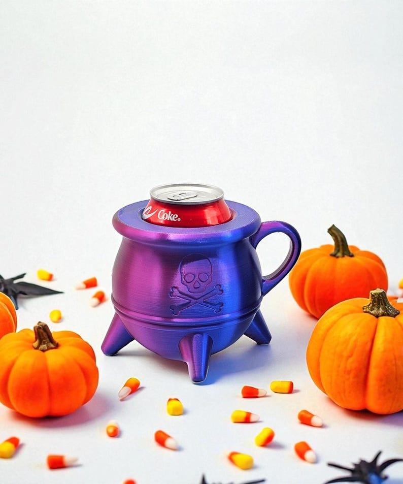 May include: A purple and blue cauldron-shaped can holder with a skull and crossbones design, holding a red Coke can. The Halloween-themed holder is surrounded by orange pumpkins and candy corn.