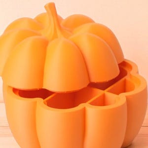 May include: An orange pumpkin-shaped container with a removable lid. The container is divided into four sections. The pumpkin has a stem and is made of a solid, smooth material. The container is likely used for storage or decoration.