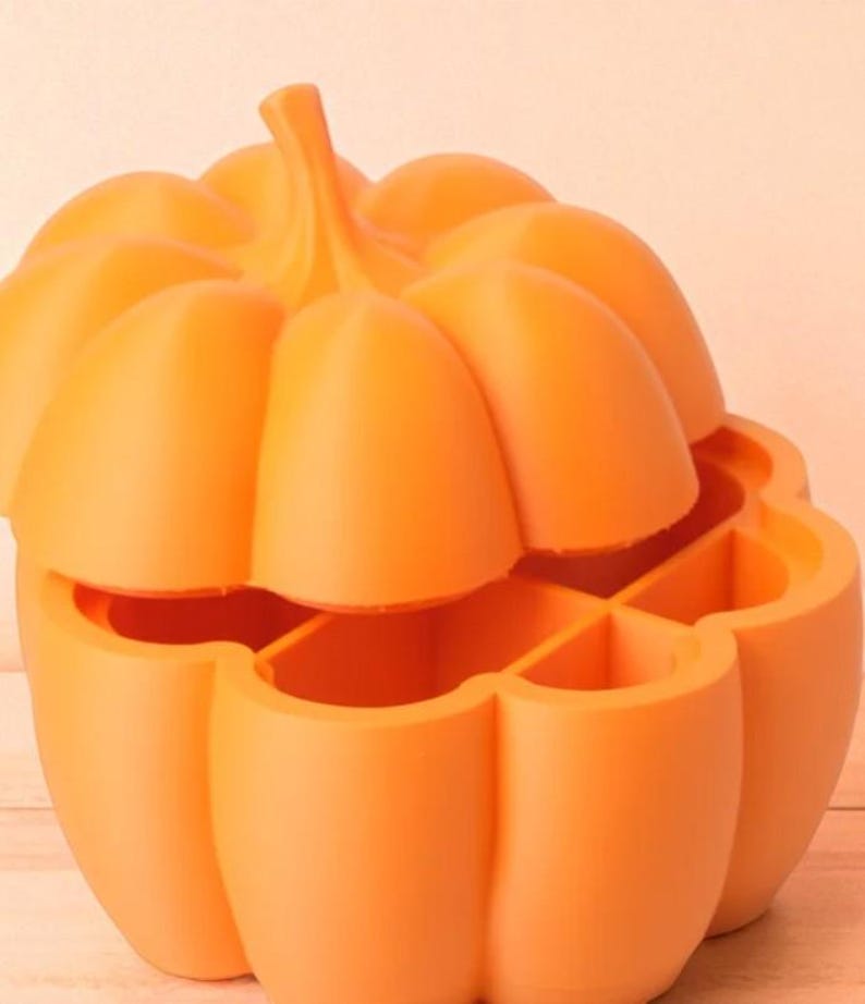 May include: An orange pumpkin-shaped container with a removable lid. The container is divided into four sections. The pumpkin has a stem and is made of a solid, smooth material. The container is likely used for storage or decoration.