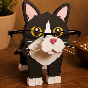 May include: A black and white cat-shaped eyeglasses holder with a pink nose and yellow eyes. The holder has a white bib and paws, and is wearing a pair of black-framed glasses. The cat is standing on a wooden surface.