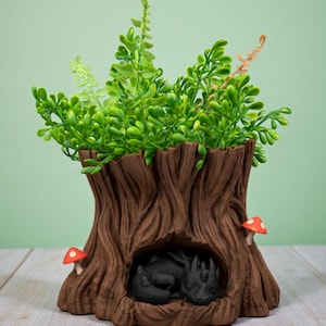 Op de afbeelding: Een bruine boomstamvormige plantenbak met groene bladeren. Twee kleine rood-witte paddenstoeldecoraties bevinden zich aan de buitenkant. Een zwart drakenfiguur nestelt zich in de holte van de boomstam.