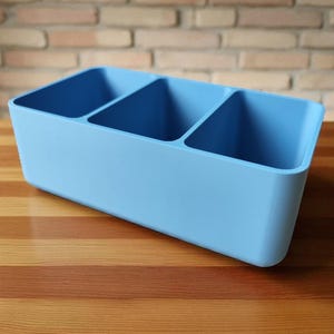 Puede incluir: Un organizador rectangular azul claro con tres compartimentos. El organizador est&aacute; hecho de un material s&oacute;lido y se encuentra sobre una superficie de madera. El fondo presenta una pared de ladrillo.
