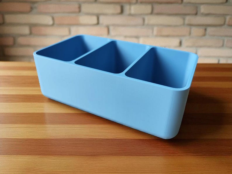Puede incluir: Un organizador rectangular azul claro con tres compartimentos. El organizador est&aacute; hecho de un material s&oacute;lido y se encuentra sobre una superficie de madera. El fondo presenta una pared de ladrillo.