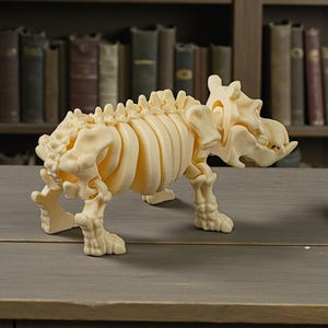 May include: A cream-colored, 3D-printed model of a prehistoric animal skeleton, possibly a dinosaur or similar creature. The model is detailed, showing ribs, spine, and skull. It stands on a wooden surface, with a blurred bookshelf background.