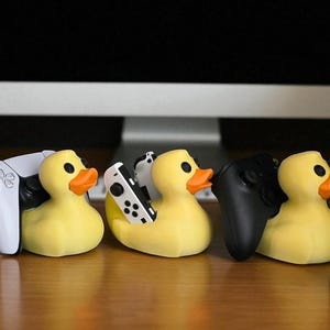 May include: Yellow rubber duck-shaped game controller holders. The ducks have orange beaks and black eyes. One duck holds a white and black game controller, another holds a black controller, and another holds a white game controller.