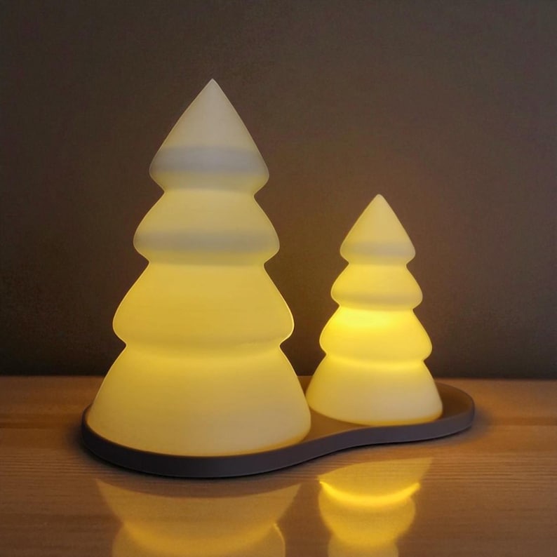May include: Two Christmas tree-shaped decorative lights. The larger tree is approximately 6 inches tall, and the smaller one is about 4 inches tall. Both are white and emit a warm, yellow glow. They sit on a dark brown, curved base, creating a festive ambiance.