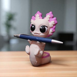 Könnte beinhalten: Eine skurrile Axolotl-Figur in Hellrosa und Lila hält einen blauen Stift. Die Figur hat große schwarze Augen und ist so positioniert, als würde sie schreiben. Der Stift hat eine Standardgröße. Das Axolotl ist ein Schreibtischzubehör.