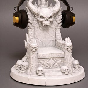 Puede incluir: Escultura de trono blanco con diseño de calavera, adornada con cuernos y una corona, con auriculares negros y dorados. El trono está rodeado de calaveras más pequeñas, creando un tema gótico o de fantasía.