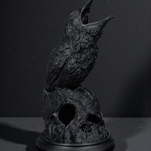 May include: A black raven statue perched atop a skull. The raven has an open beak, and the skull has a hollow eye socket. The statue is on a round base. The sculpture is a gothic or macabre decorative item.