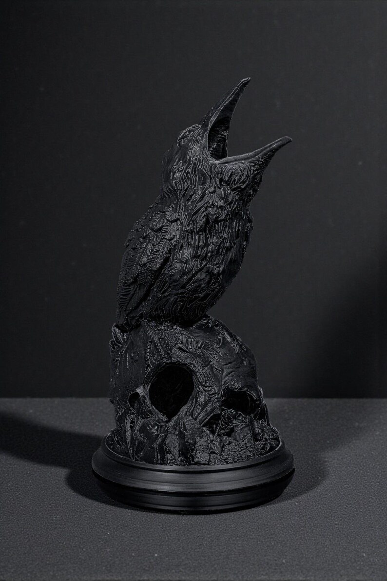 May include: A black raven statue perched atop a skull. The raven has an open beak, and the skull has a hollow eye socket. The statue is on a round base. The sculpture is a gothic or macabre decorative item.