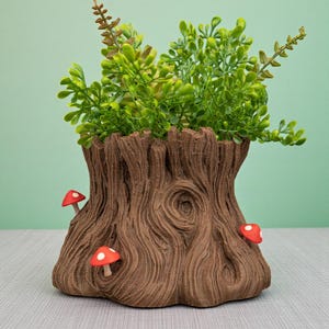Op de afbeelding: Een bruine boomstronk-vormige plantenbak met een getextureerd oppervlak, met drie rode en witte paddenstoelaccenten. De plantenbak is gevuld met groen gebladerte. De achtergrond is effen lichtgroen.