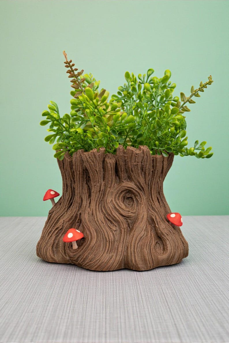 Op de afbeelding: Een bruine boomstronk-vormige plantenbak met een getextureerd oppervlak, met drie rode en witte paddenstoelaccenten. De plantenbak is gevuld met groen gebladerte. De achtergrond is effen lichtgroen.
