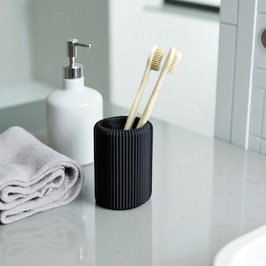 May include: A black, ribbed toothbrush holder containing two bamboo toothbrushes. A white soap dispenser and a folded grey towel are also present. The items are arranged on a reflective, light-coloured surface, suggesting a bathroom setting.