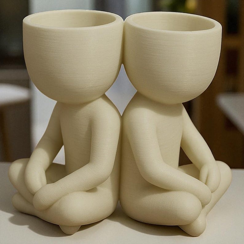 May include: Two off-white ceramic planters shaped like seated figures with large, rounded heads. The figures are in a cross-legged position, with their arms resting on their knees. The planters are designed to hold small plants or succulents.