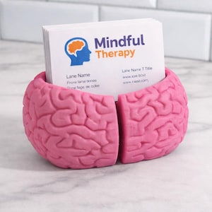 May include: A pink brain-shaped business card holder containing white cards. The cards display the text "Mindful Therapy" and contact details. The holder is made of a solid material and rests on a marble surface.