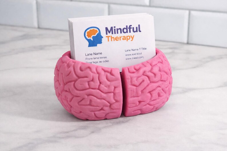 May include: A pink brain-shaped business card holder containing white cards. The cards display the text "Mindful Therapy" and contact details. The holder is made of a solid material and rests on a marble surface.