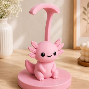 May include: A pink axolotl-shaped phone stand. The stand features a cartoon axolotl with black eyes and a smiling mouth, sitting on a round base. A curved support extends upwards from the base. The stand is made of a solid pink material.