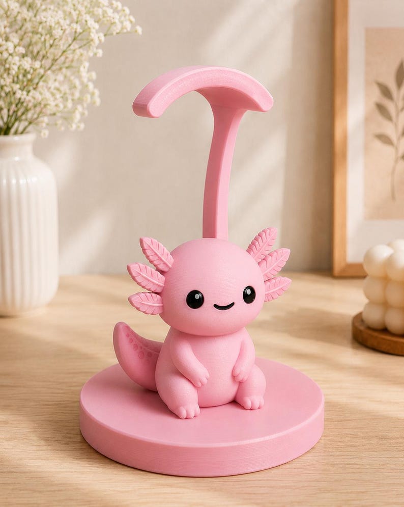 May include: A pink axolotl-shaped phone stand. The stand features a cartoon axolotl with black eyes and a smiling mouth, sitting on a round base. A curved support extends upwards from the base. The stand is made of a solid pink material.