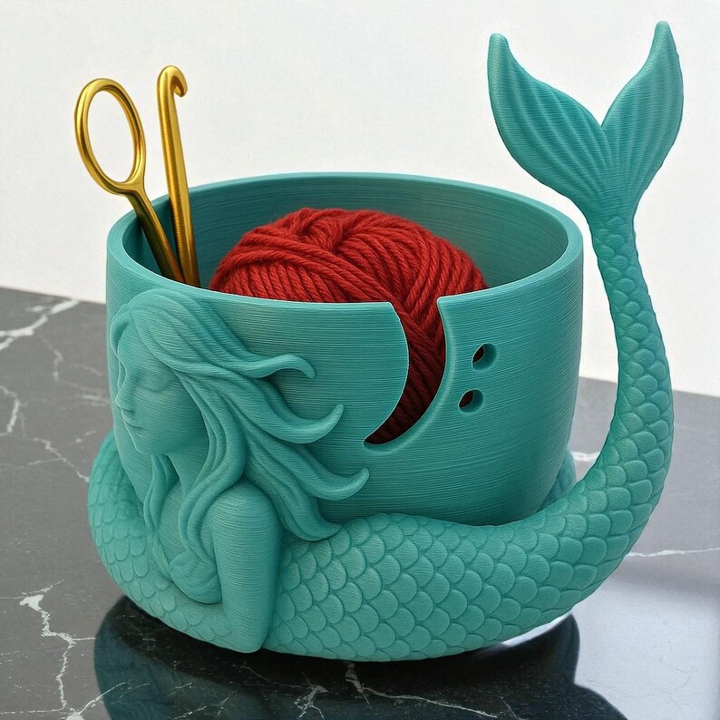 May include: A teal mermaid-shaped yarn bowl with a red yarn ball inside. The bowl has a mermaid tail and a sculpted mermaid figure on the side. Gold crochet hook and scissors are inside the bowl. The bowl is designed for holding yarn while crocheting or knitting.