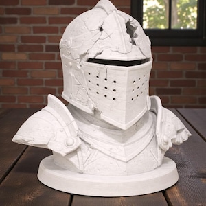 Puede incluir: Busto blanco de un casco y hombreras de caballero, con aspecto envejecido. El casco tiene una visera con peque&ntilde;os agujeros y est&aacute; montado sobre una base circular. El busto se exhibe sobre una superficie de madera oscura.