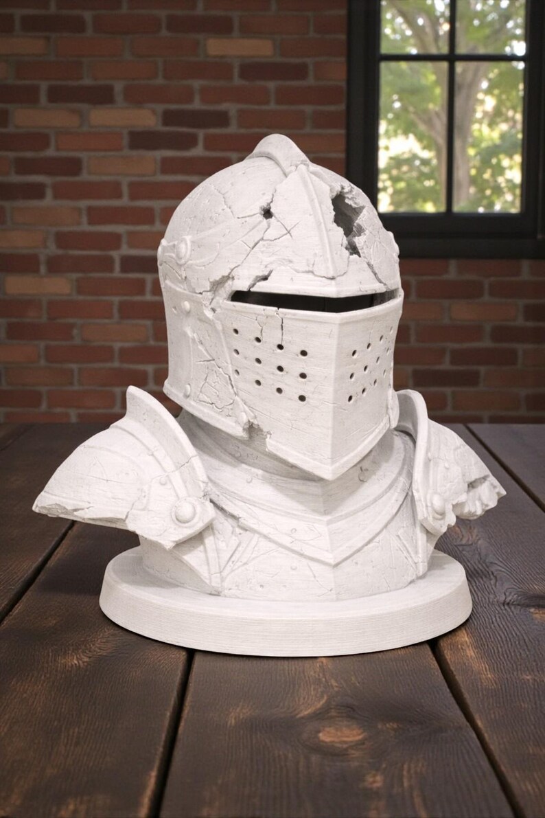 Puede incluir: Busto blanco de un casco y hombreras de caballero, con aspecto envejecido. El casco tiene una visera con peque&ntilde;os agujeros y est&aacute; montado sobre una base circular. El busto se exhibe sobre una superficie de madera oscura.