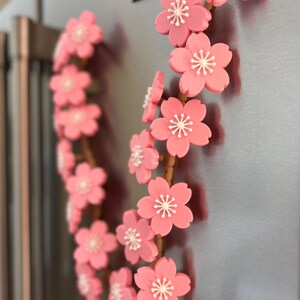 May include: A decorative floral wreath featuring pink cherry blossoms with white centers. The blossoms are arranged on a brown stem, creating a delicate and natural aesthetic. The wreath is attached to a metallic surface.