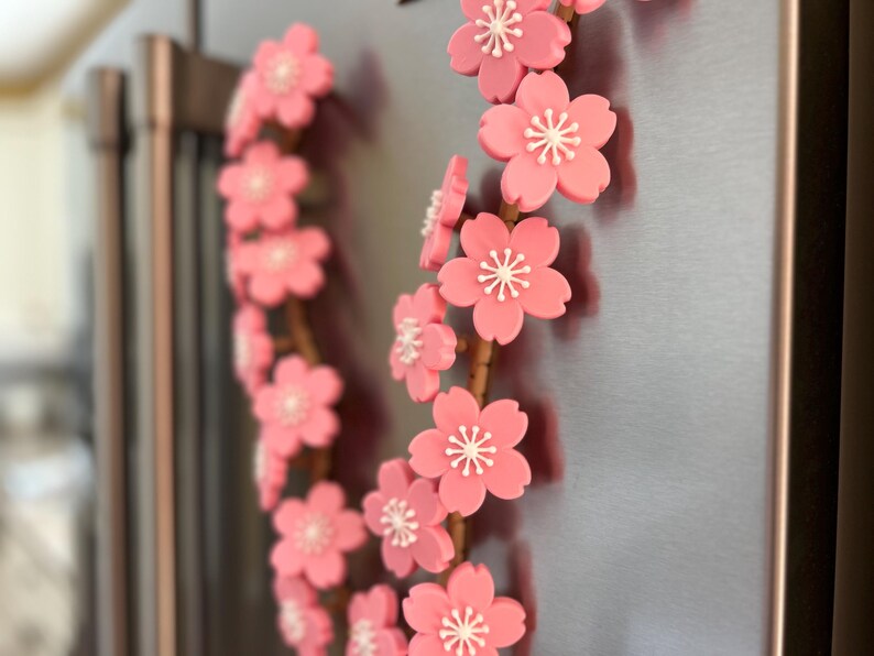 May include: A decorative floral wreath featuring pink cherry blossoms with white centers. The blossoms are arranged on a brown stem, creating a delicate and natural aesthetic. The wreath is attached to a metallic surface.