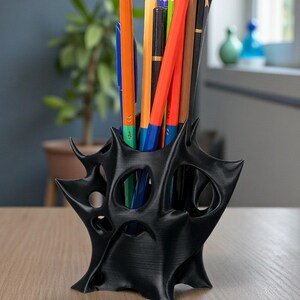 May include: Black, abstract-shaped desk organizer holding a variety of colored pencils and a blue pen. The organizer has a unique, open design with multiple holes. The pencils are orange, red, brown, and black.