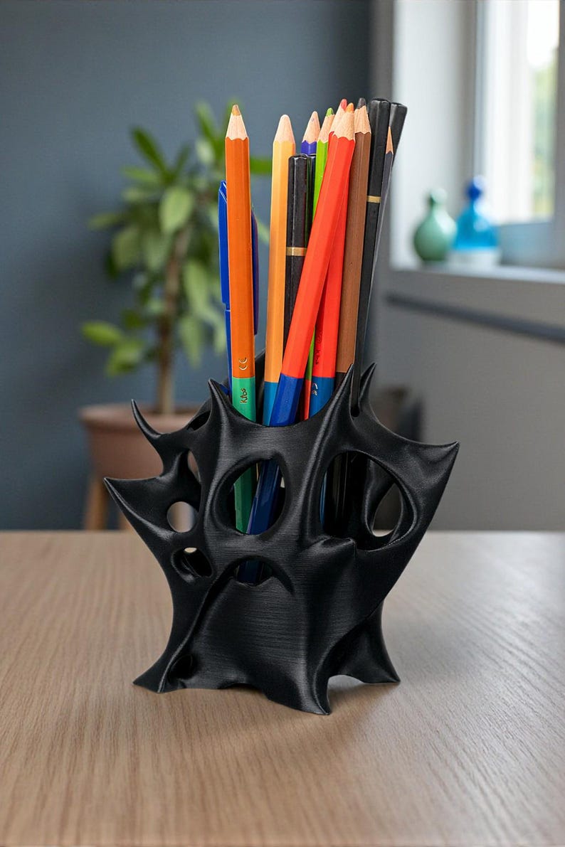 May include: Black, abstract-shaped desk organizer holding a variety of colored pencils and a blue pen. The organizer has a unique, open design with multiple holes. The pencils are orange, red, brown, and black.
