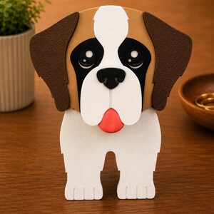 May include: A decorative, three-dimensional Saint Bernard dog figurine. The dog has a white body, brown ears, and a brown and white head. The dog's face features black eyes, a black nose, and a red tongue. The figurine is standing on a wooden surface.