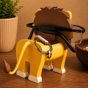 May include: A whimsical lion-shaped eyeglasses holder in yellow, brown, and white. The lion's mane and tail tip are brown, with white accents on the paws. Black eyeglasses rest on the lion's head, and a key ring hangs from the back.