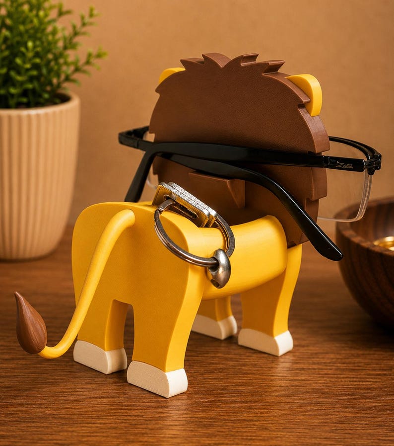 May include: A whimsical lion-shaped eyeglasses holder in yellow, brown, and white. The lion's mane and tail tip are brown, with white accents on the paws. Black eyeglasses rest on the lion's head, and a key ring hangs from the back.