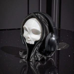 May include: A white skull-shaped headphone stand wearing a black hooded cloak, with black wireless headphones resting on the stand. The stand is on a black, reflective surface.
