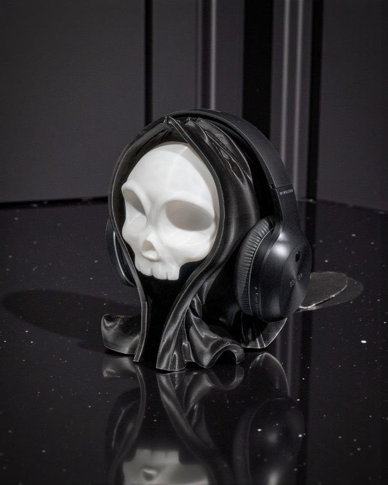 May include: A white skull-shaped headphone stand wearing a black hooded cloak, with black wireless headphones resting on the stand. The stand is on a black, reflective surface.
