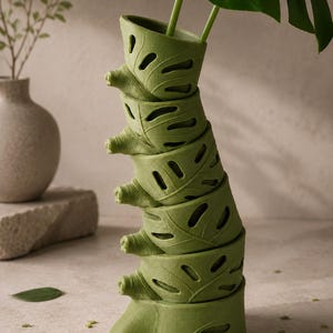 Puede incluir: Una jardinera verde apilada con forma de columna vertebral, con recortes en forma de hoja. La jardinera contiene una gran hoja verde y tallos. El dise&ntilde;o es moderno y org&aacute;nico, adecuado para plantas de interior. La jardinera mide aproximadamente 30 cm de altura.