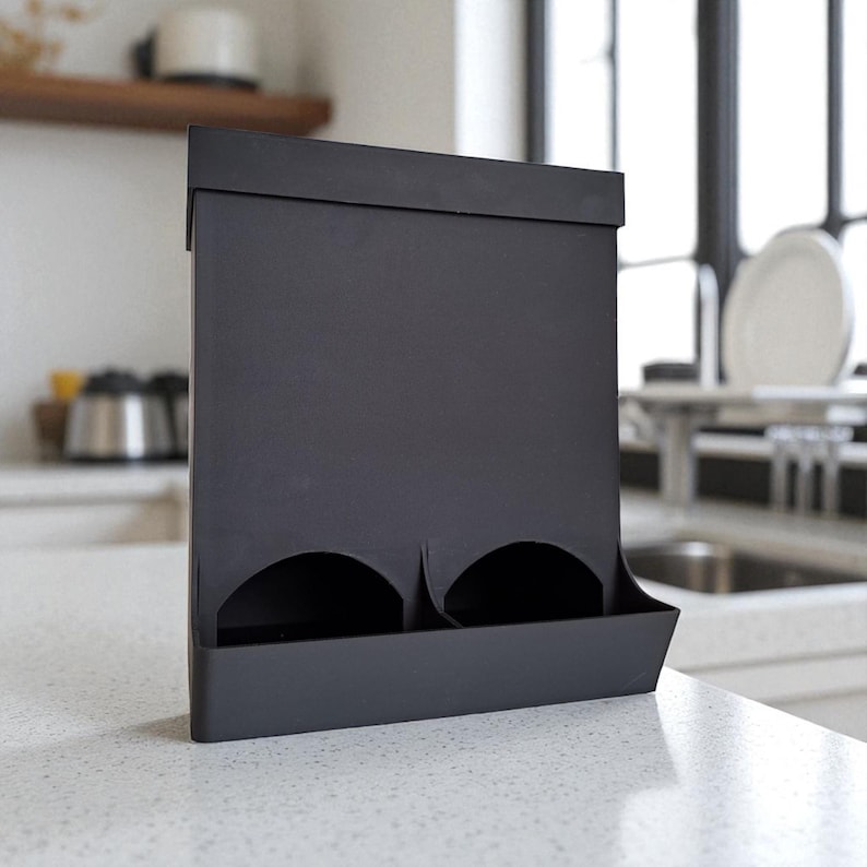 May include: A matte black kitchen organizer with two circular openings and a tray at the bottom. The organizer is made of a solid material and sits on a white countertop. The design is modern and minimalist.