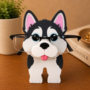 May include: A whimsical eyeglass holder shaped like a cartoon husky dog. The holder is black, white, and pink, with blue eyes and a pink tongue. The glasses are held in place by the dog's nose and mouth. A fun and functional desk accessory.