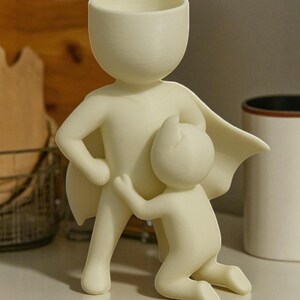 Op de afbeelding: Een cr&egrave;mekleurige decoratieve plantenbak in de vorm van een superheld met een kind dat zich aan zijn been vastklampt. De superheld heeft een cape en een grote kop die de opening van de plantenbak vormt. De sculptuur staat op een wit oppervlak.