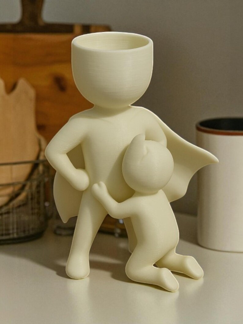 Op de afbeelding: Een cr&egrave;mekleurige decoratieve plantenbak in de vorm van een superheld met een kind dat zich aan zijn been vastklampt. De superheld heeft een cape en een grote kop die de opening van de plantenbak vormt. De sculptuur staat op een wit oppervlak.