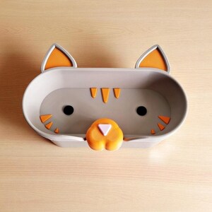 May include: A grey cat-shaped tray with orange detailing. The tray has a cat face design, including black eyes, orange ears, and orange triangular features. The nose is orange with a white triangle. The tray is designed for small items.