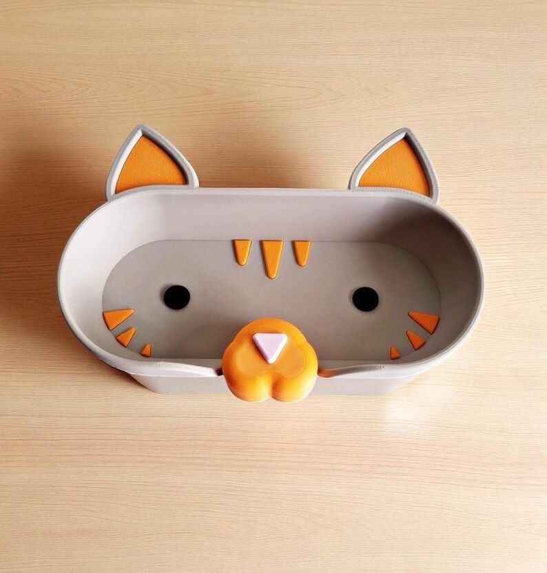 May include: A grey cat-shaped tray with orange detailing. The tray has a cat face design, including black eyes, orange ears, and orange triangular features. The nose is orange with a white triangle. The tray is designed for small items.