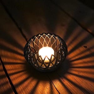 May include: A lit candle in a decorative, dark metal holder with a lattice design. The candle's warm glow casts intricate shadows on the wooden surface, creating a cozy ambiance. The candle holder is round and has a geometric pattern.
