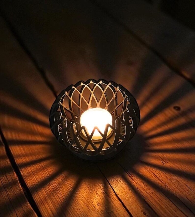 May include: A lit candle in a decorative, dark metal holder with a lattice design. The candle's warm glow casts intricate shadows on the wooden surface, creating a cozy ambiance. The candle holder is round and has a geometric pattern.