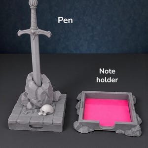 May include: A gray pen holder shaped like a sword in a stone, with a small skull and a matching note holder with a pink pad. The word "Pen" is written above the pen holder and "Note holder" above the note holder.