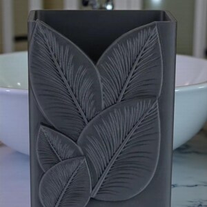 May include: A dark gray rectangular bathroom organizer with a leaf design. The leaves are raised and textured, adding dimension to the container. The organizer is likely made of a solid material and is designed to hold items like toothbrushes or makeup brushes.