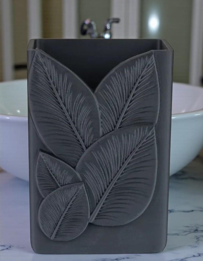 May include: A dark gray rectangular bathroom organizer with a leaf design. The leaves are raised and textured, adding dimension to the container. The organizer is likely made of a solid material and is designed to hold items like toothbrushes or makeup brushes.