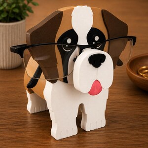 May include: A wooden dog-shaped eyeglass holder. The dog has brown and white markings, black eyes, and a pink tongue. It wears black-framed glasses. The holder is designed to hold eyeglasses.